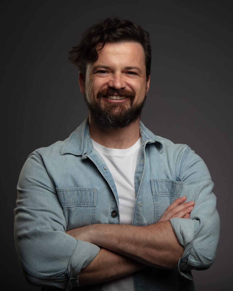 Marc Owen Jones, smiling, arms folded, in a denim shirt against a dark grey backdrop